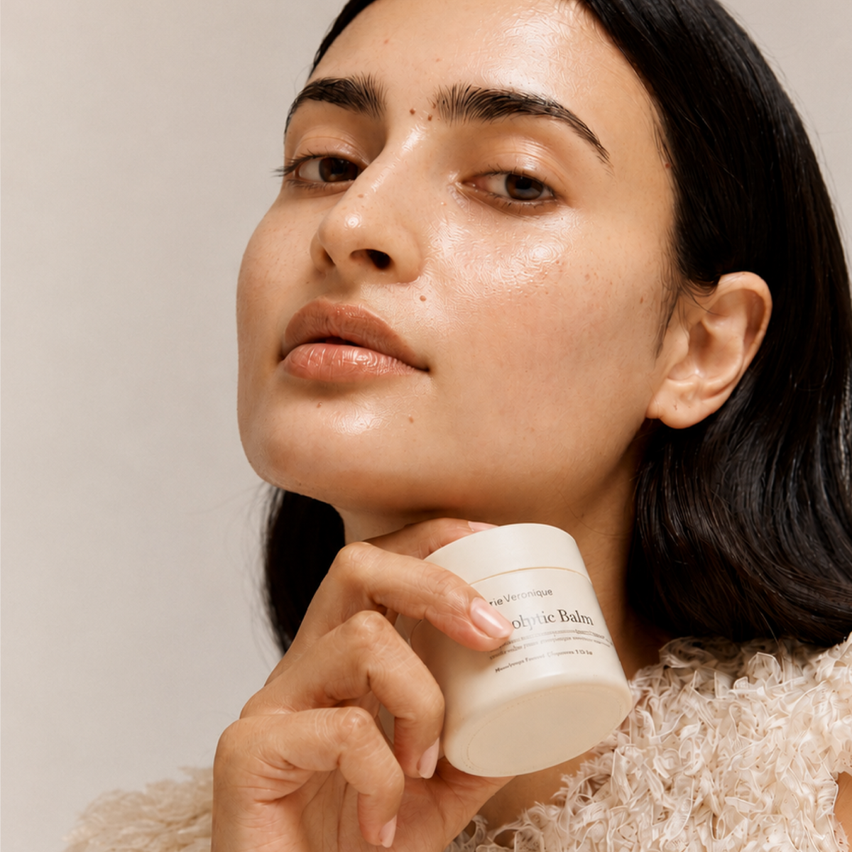 A close-up lifestyle shot of a woman with a glowing, hydrated complexion holding a white jar of 'Marie Veronique' Senolytic Balm against a soft beige background.