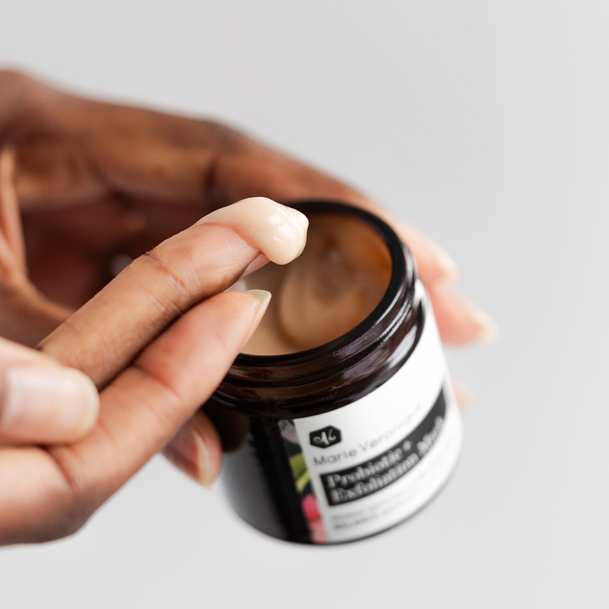 Hand holding jar and other hand taking a scoop of product to show texture of Marie Veronique Probiotic + Exfoliation Mask 1.7 oz / 50ml. Restore skin’s natural MICROFLORAL BALANCE and brighten with ENZYMES. Microbiome-friendly